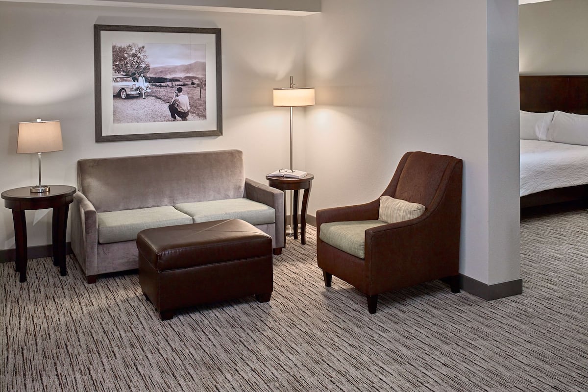A hotel room with a sofa, armchair, and ottoman, a lamp, a framed picture on the wall, and a bed with white linens.
