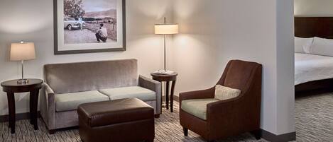 Een hotelkamer met een bank, fauteuil, een poef, een lamp, een ingelijste foto aan de muur en een bed met witte lakens.