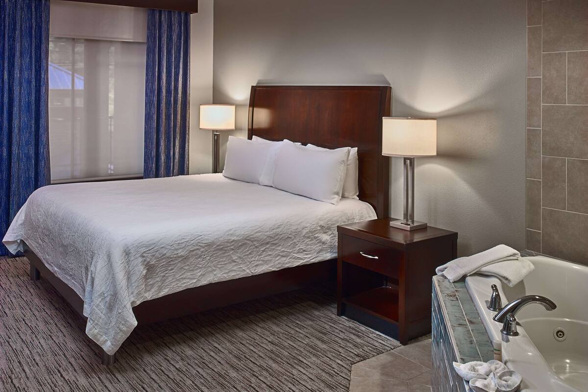 A hotel room with a large bed, two bedside lamps, a wooden nightstand, a bathroom with a bathtub, and a window with blue curtains.