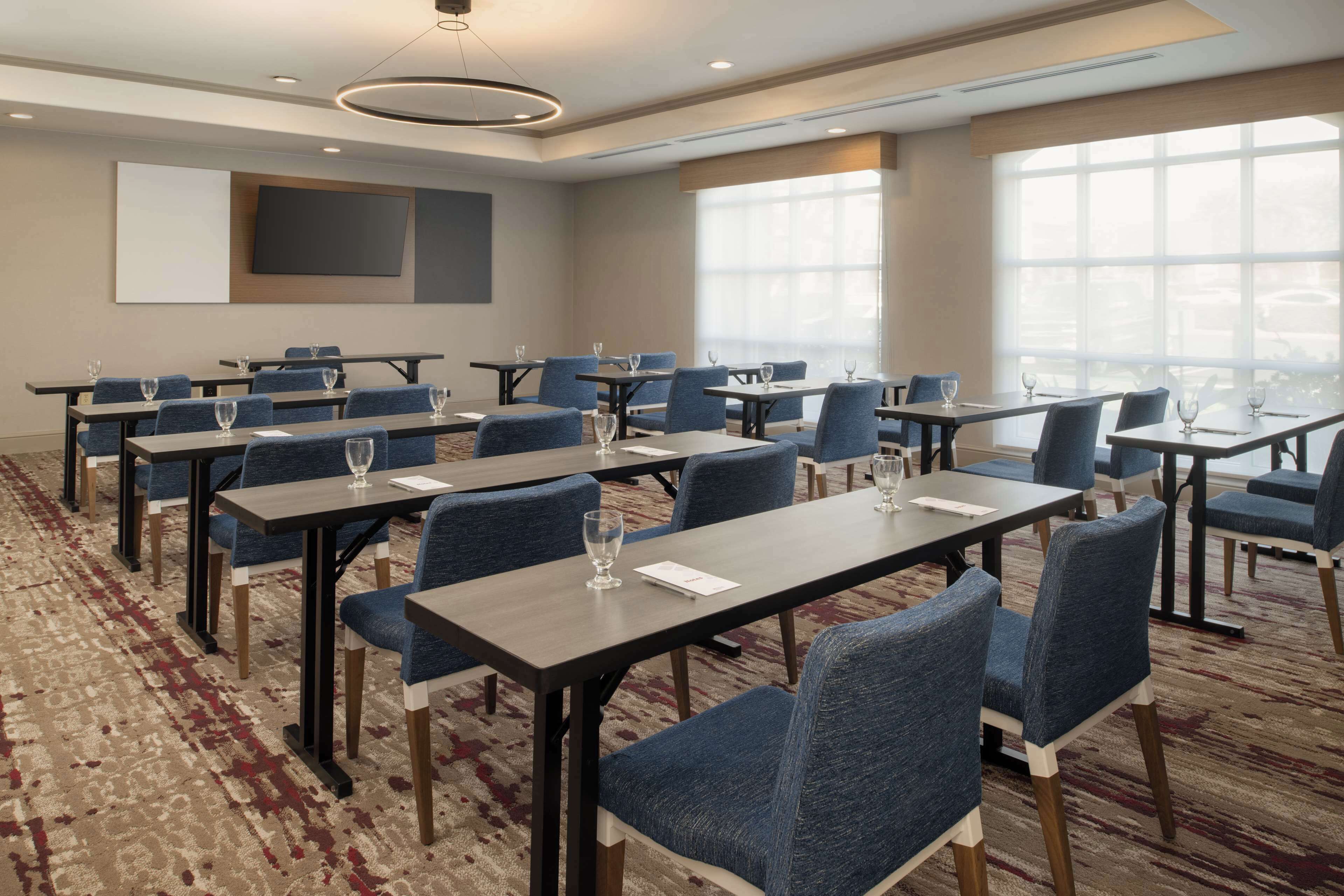 A conference room with multiple tables and chairs, large windows, and a carpeted floor.