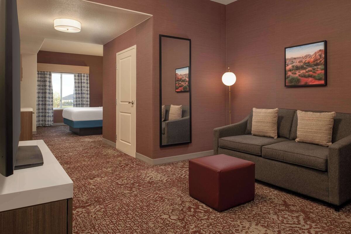 Habitación de hotel con cama, sofá, reposapiés rojo, televisor sobre un mueble, espejo grande y dos cuadros en la pared.