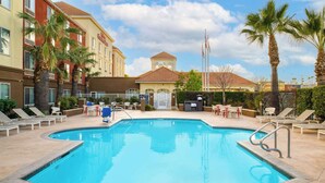 Ein Swimmingpool mit Liegestühlen und Palmen, umgeben von einem Hotelgebäude mit Ziegeldach.