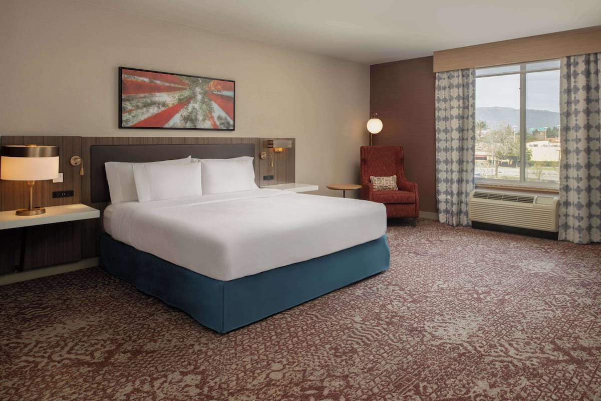 Habitación de hotel con una cama grande, un escritorio, una silla, un ventanal con cortinas y una obra de arte en la pared.