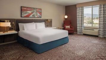 A hotel room with a large bed, a desk, a chair, a window with curtains, and a wall-mounted artwork.