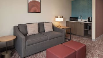 A modern hotel room with a grey sofa, a small round table, a red ottoman, a kitchenette with a microwave, coffee maker, and a sink.