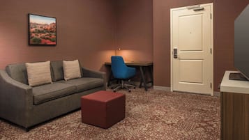 Una habitación de hotel con un sofá gris, un taburete rojo, una silla azul, un escritorio y una puerta.