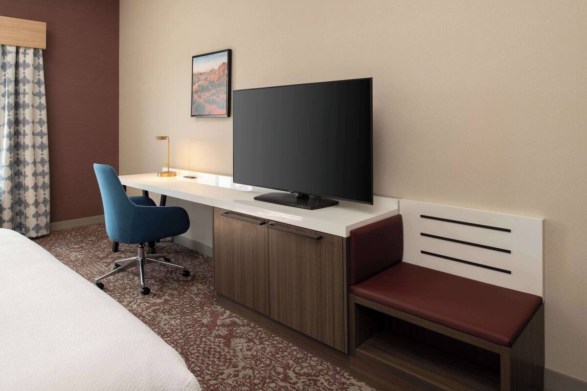 Habitación de hotel con cama, escritorio con televisor, una silla azul, un banco rojo y una alfombra estampada.