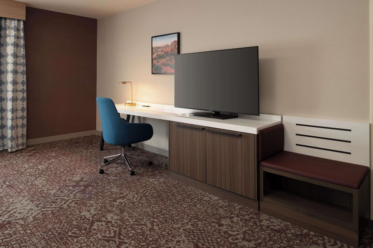 Habitación de hotel con una televisión de pantalla plana sobre un escritorio blanco, una silla de oficina azul, un armario de madera y un banco rojo.