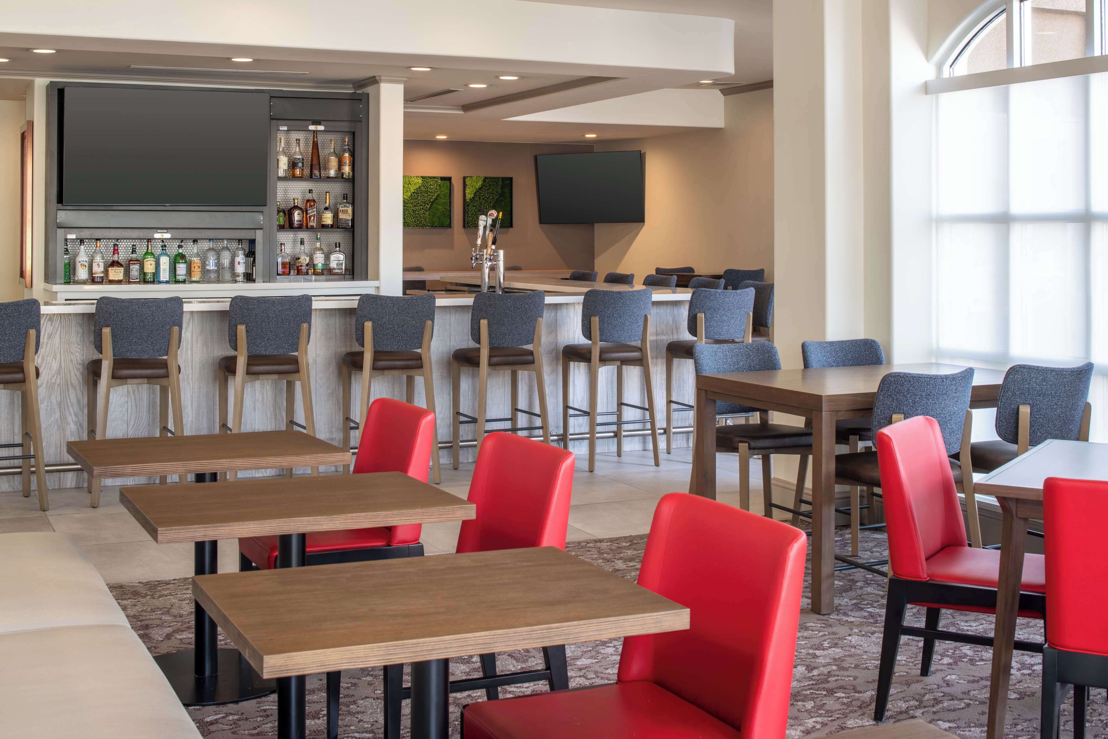 A modern bar area with a variety of liquor bottles, a flat-screen TV, and a seating area with red and grey chairs and wooden tables.