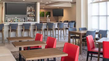 A modern bar area with a variety of liquor bottles, a flat-screen TV, and a seating area with red and grey chairs and wooden tables.