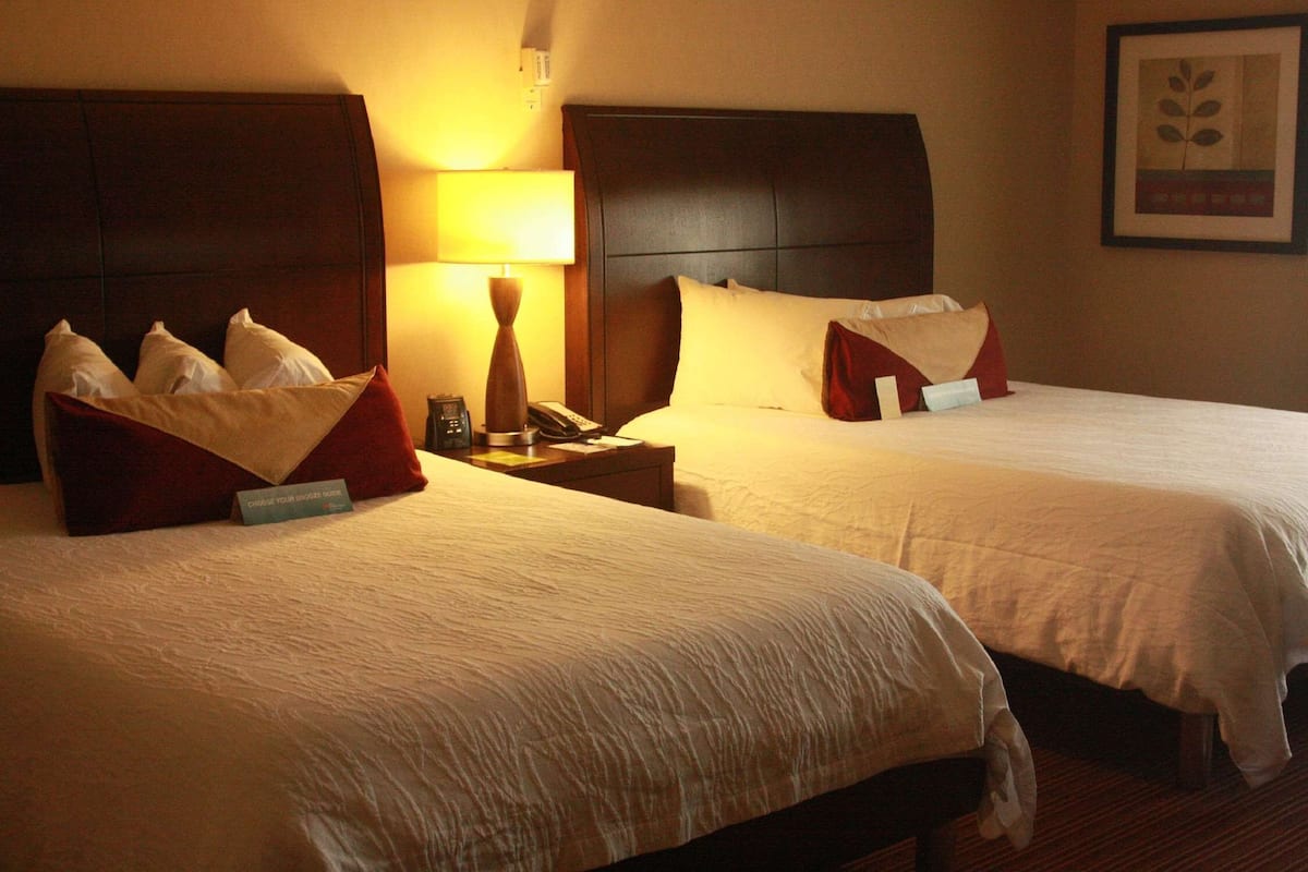 Habitación de hotel con dos camas, cada una con ropa blanca y almohadas rojas, una mesita de noche con lámpara y un cuadro en la pared.