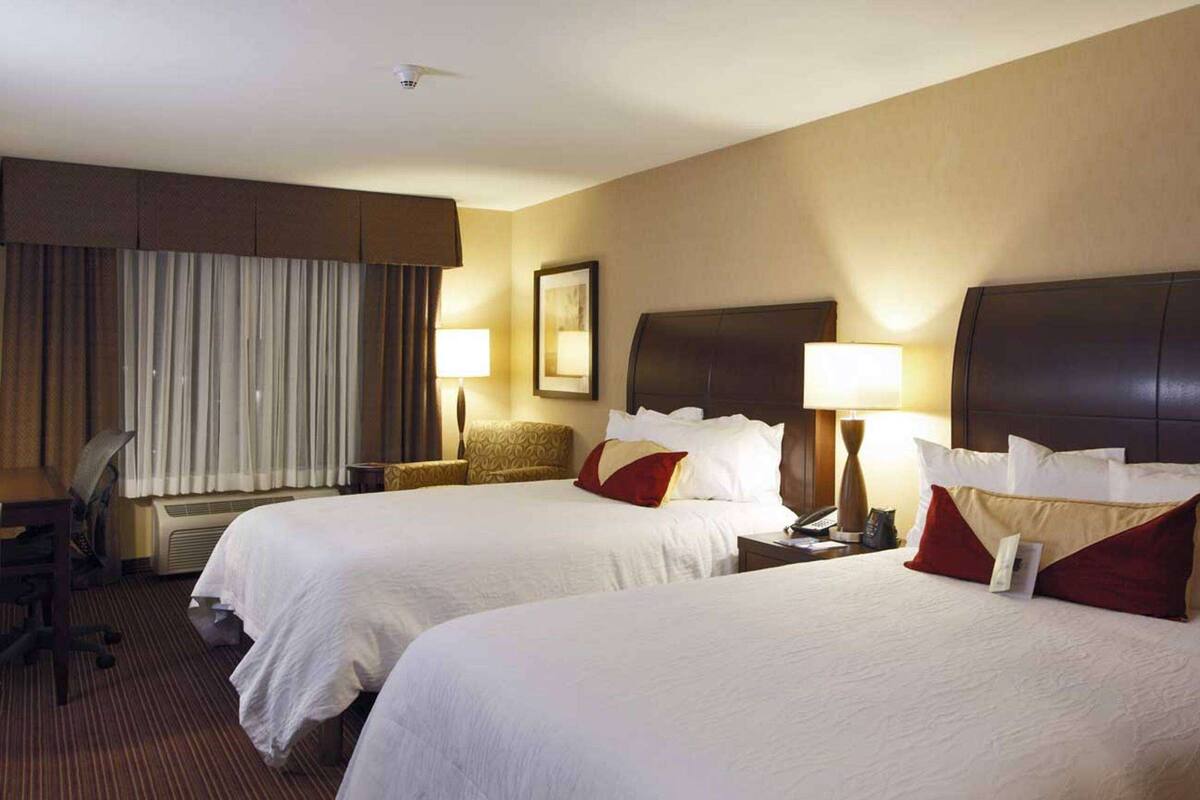 Habitación de hotel con dos camas, un escritorio, una silla y una ventana con cortinas.