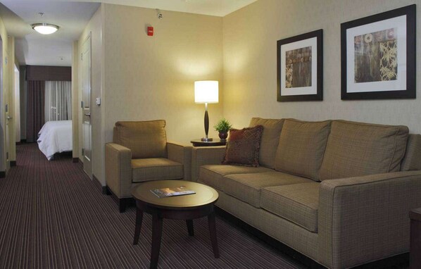 A hotel room with a sofa, armchair, and a small table with a lamp. There is a bed in the background and two framed pictures on the wall.