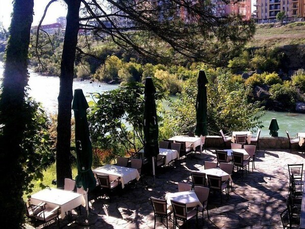 Un espacio de comedor al aire libre con mesas y sillas, rodeado de árboles y vistas a un río.