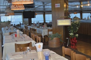 Un restaurant avec de longues tables dressées pour le service, une enseigne indiquant qu’il s’agit d’un restaurant, et un téléviseur fixé au mur.