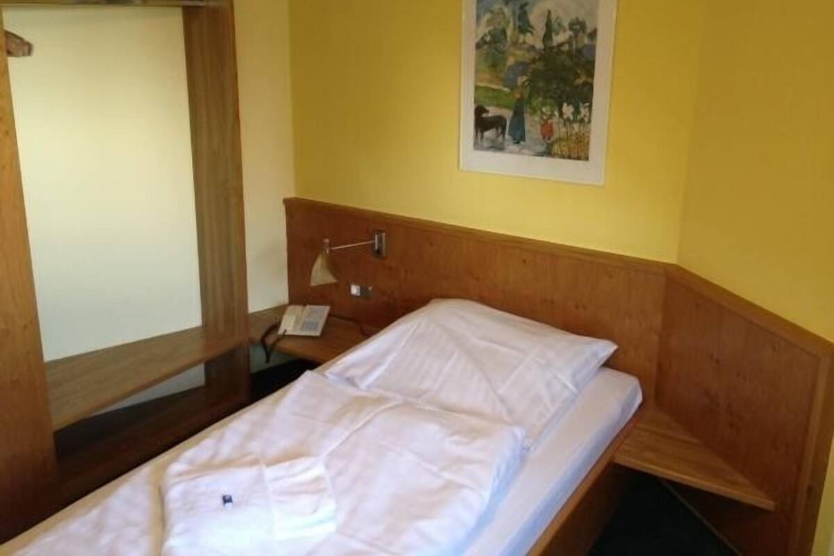 Una cama individual con cabecera de madera, una mesita de noche con lámpara y un cuadro en la pared.