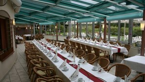 Una larga mesa de comedor dispuesta para un evento formal, rodeada de sillas de mimbre, bajo techo de cristal con vistas al jardín.