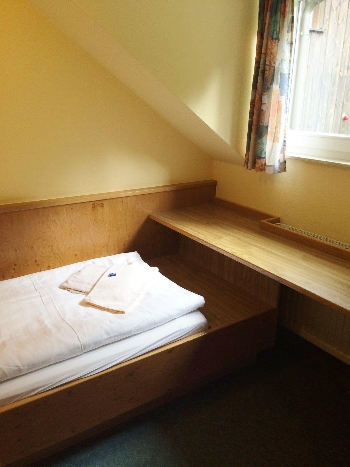 Ein kleiner, schlichter Raum mit einem Holzbett, einem kleinen Schreibtisch und einem Fenster mit Vorhängen.