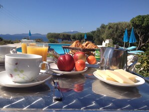 A breakfast table with croissants, apples, and orange juice, overlooking a pool and mountains. - Hôtel Céline (Coti-Chiavari)