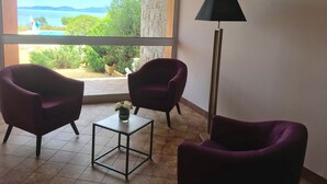 A room with two purple armchairs, a small table with a vase, and a floor lamp. A large window offers a view of greenery and a distant landscape. - Hôtel Céline (Coti-Chiavari)