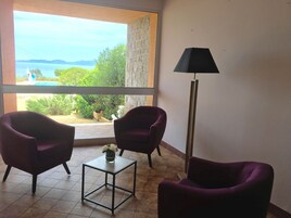 Una habitación con dos sillones morados, una pequeña mesa con un florero y una lámpara de pie. Un amplio ventanal ofrece vistas a la vegetación y un paisaje lejano.