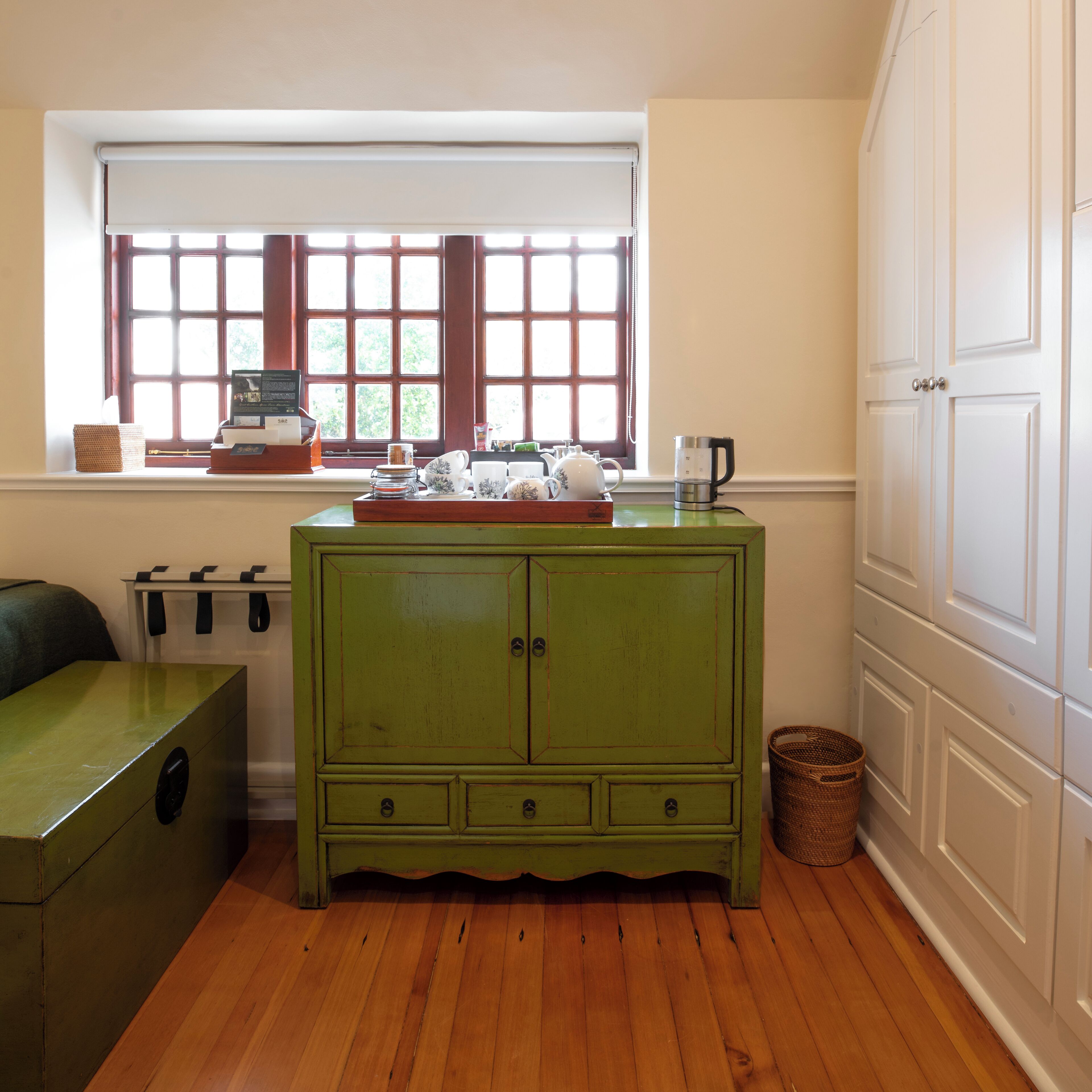 Una habitación con un aparador verde, una ventana con persianas, piso de madera y una puerta blanca.