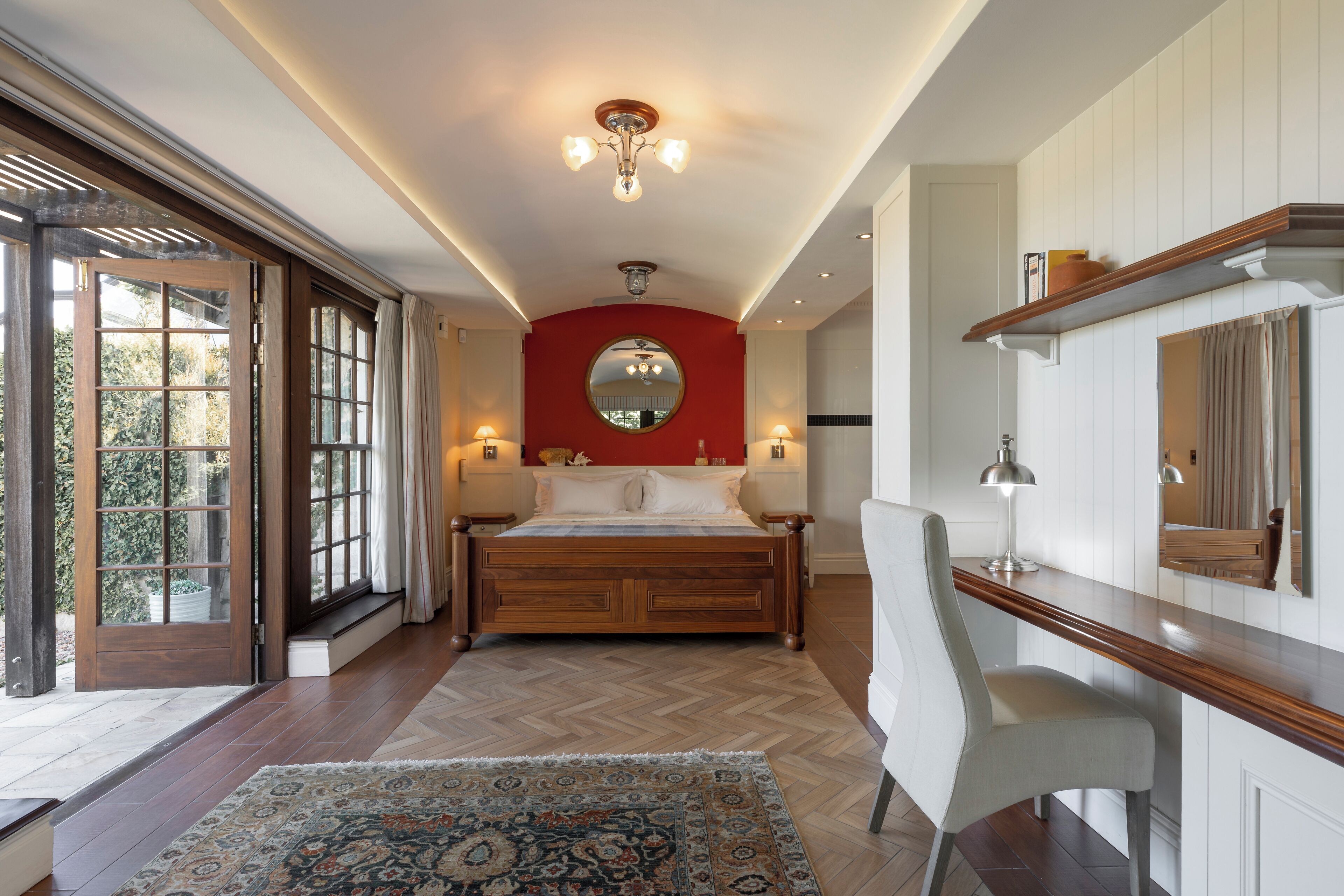 Ein Schlafzimmer mit einem großen Bett, einem Schreibtisch, einem Stuhl, einem runden Spiegel und einer roten Wand als Gestaltungselement.
