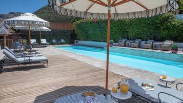 Zona junto a la piscina con sillones de descanso, una mesa con bebidas y sombrillas.