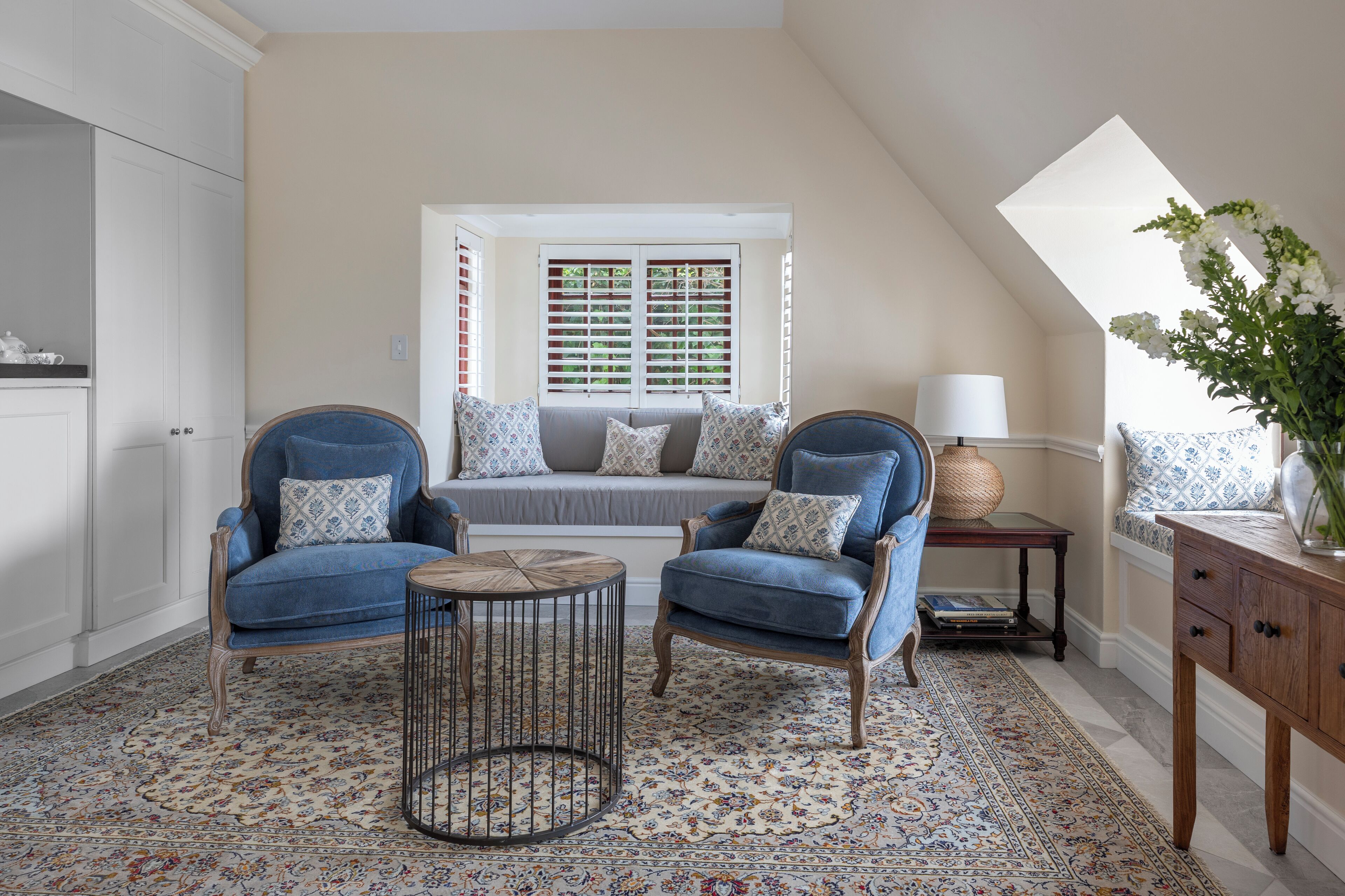 A cozy living room with a high ceiling, a large window with shutters, a blue sofa set, a small round coffee table, and a decorative lamp.
