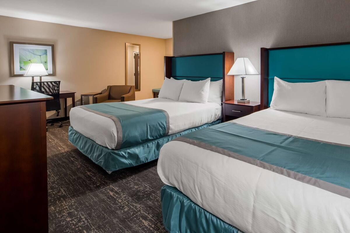 A hotel room with two beds, a desk, a chair, a lamp, and a framed picture on the wall.