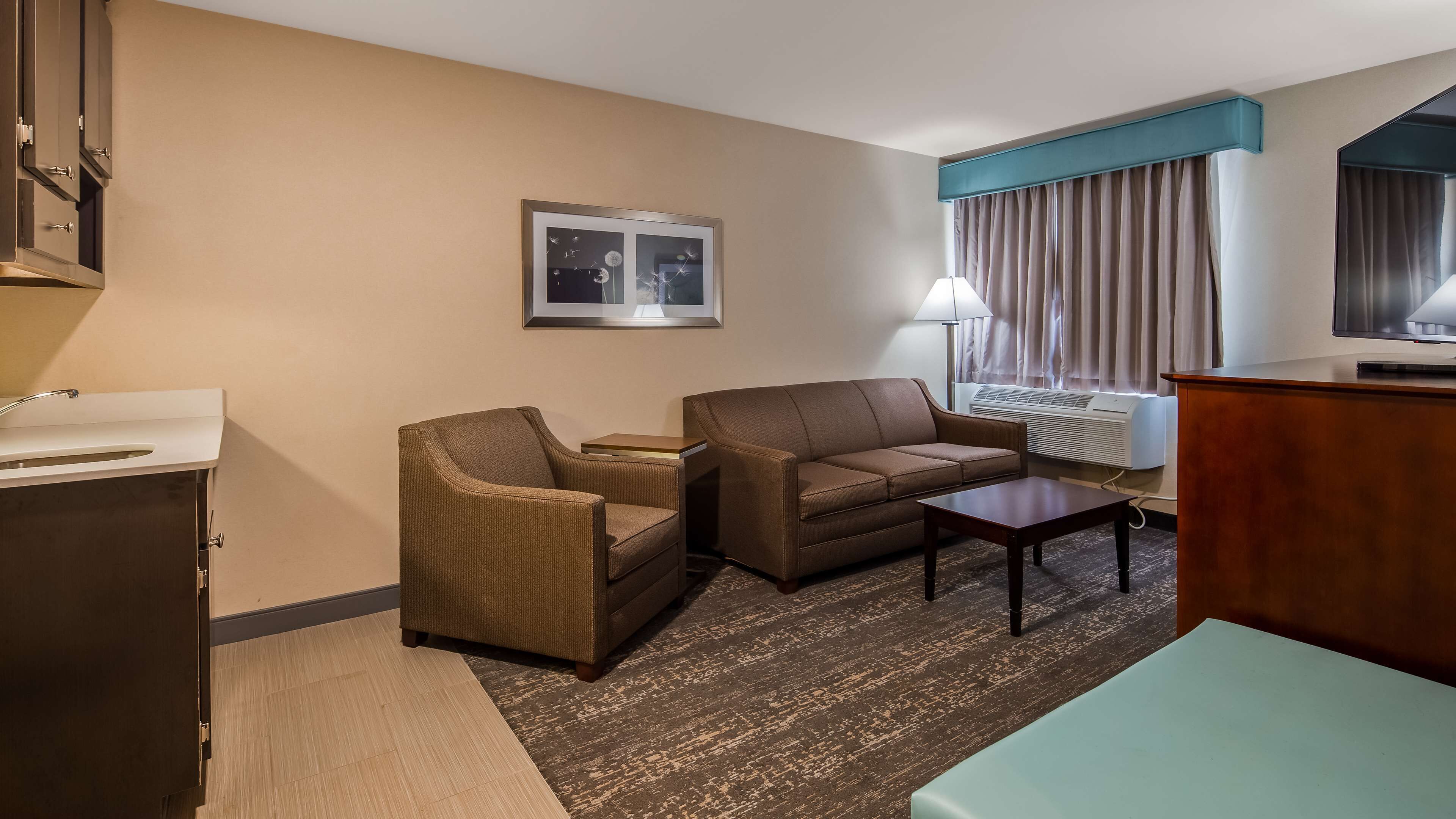 A hotel room with a sofa, armchair, small table, and a desk with a lamp.