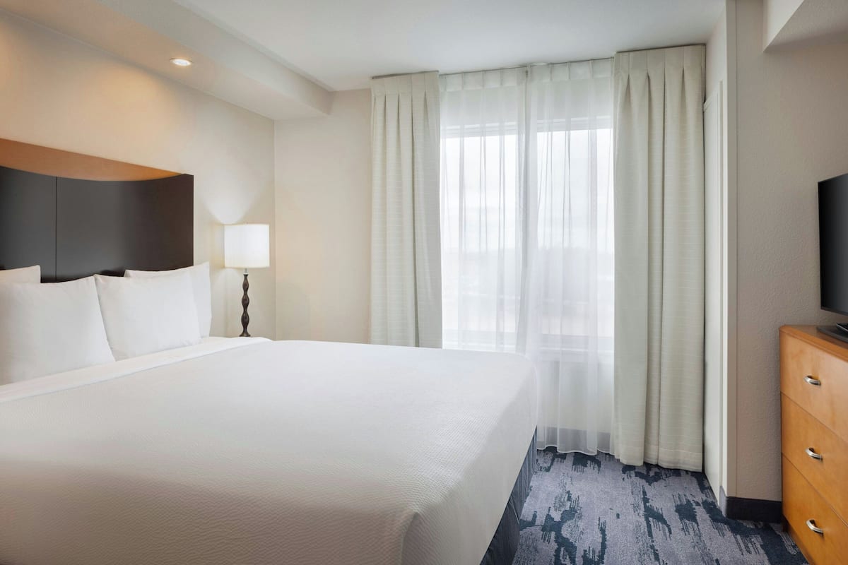 A hotel room with a large bed, a TV, a wooden dresser, and a window with sheer curtains.