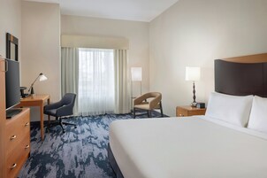 Quarto de hotel com uma cama grande, uma escrivaninha com cadeira, televisão, abajur e janela com cortinas.