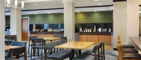 A modern dining area with tables and chairs, a kitchen in the background, and a green wall.
