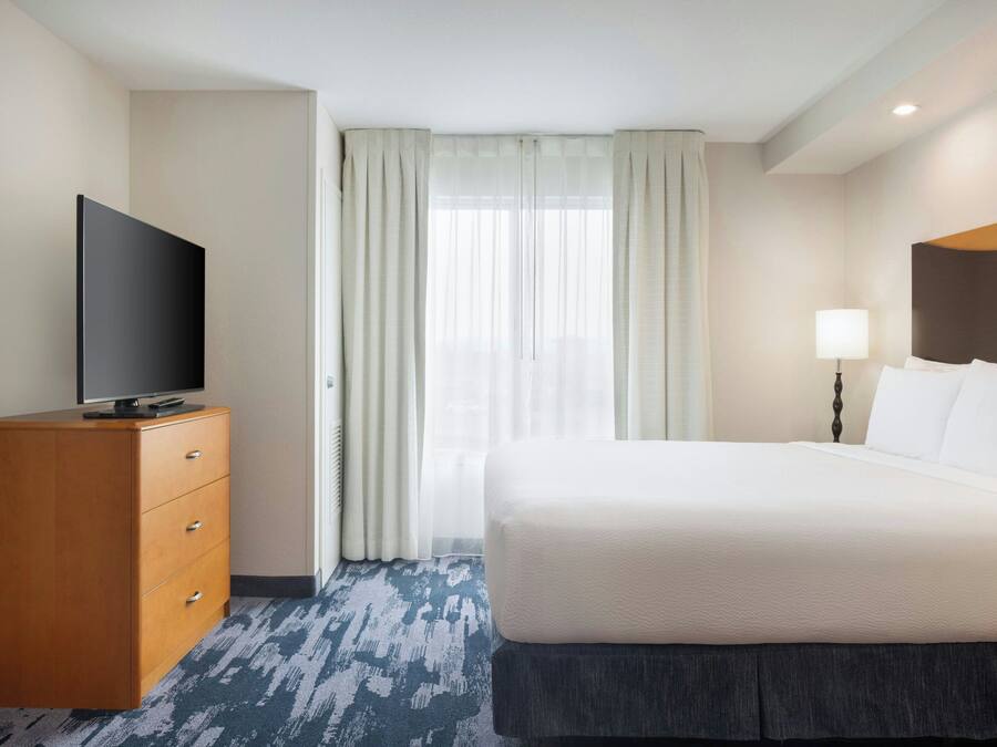 Quarto de hotel com uma cama, uma TV sobre uma cômoda de madeira e uma janela com cortinas.