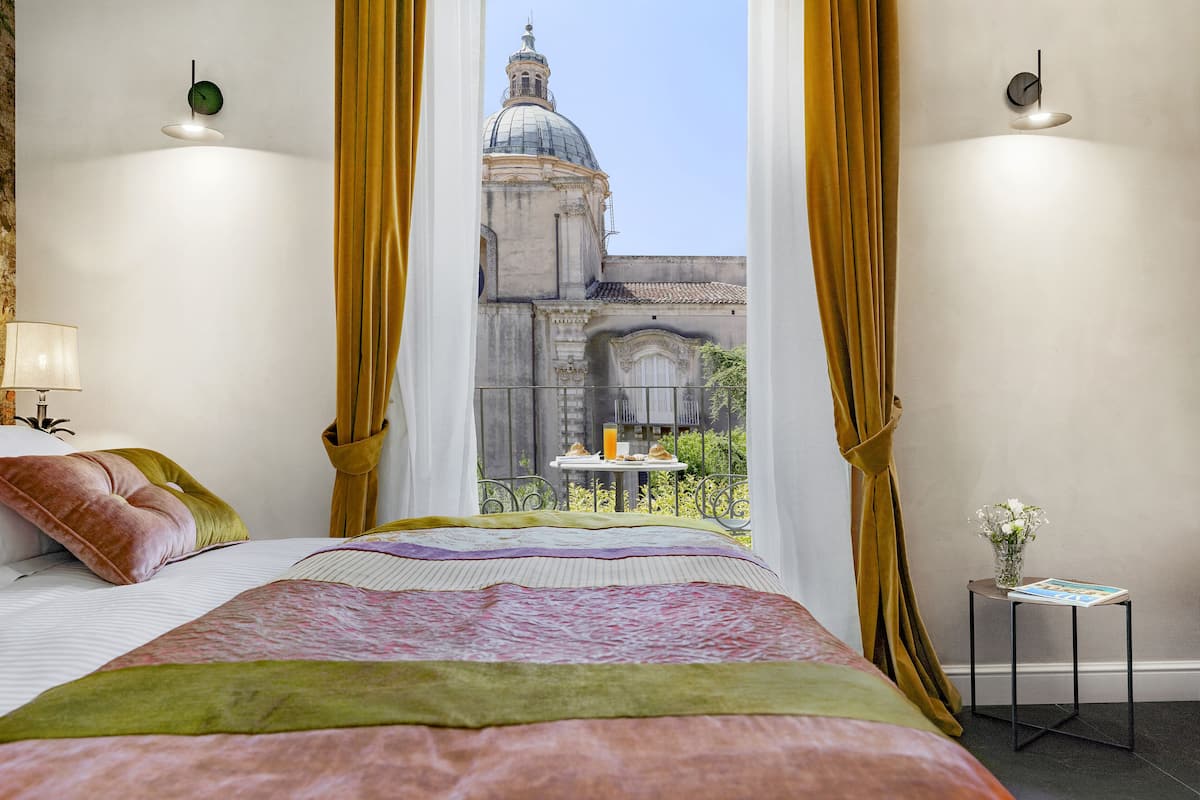 A hotel room with a large bed, a view of a historic building, a small table with a vase of flowers, and a lamp.