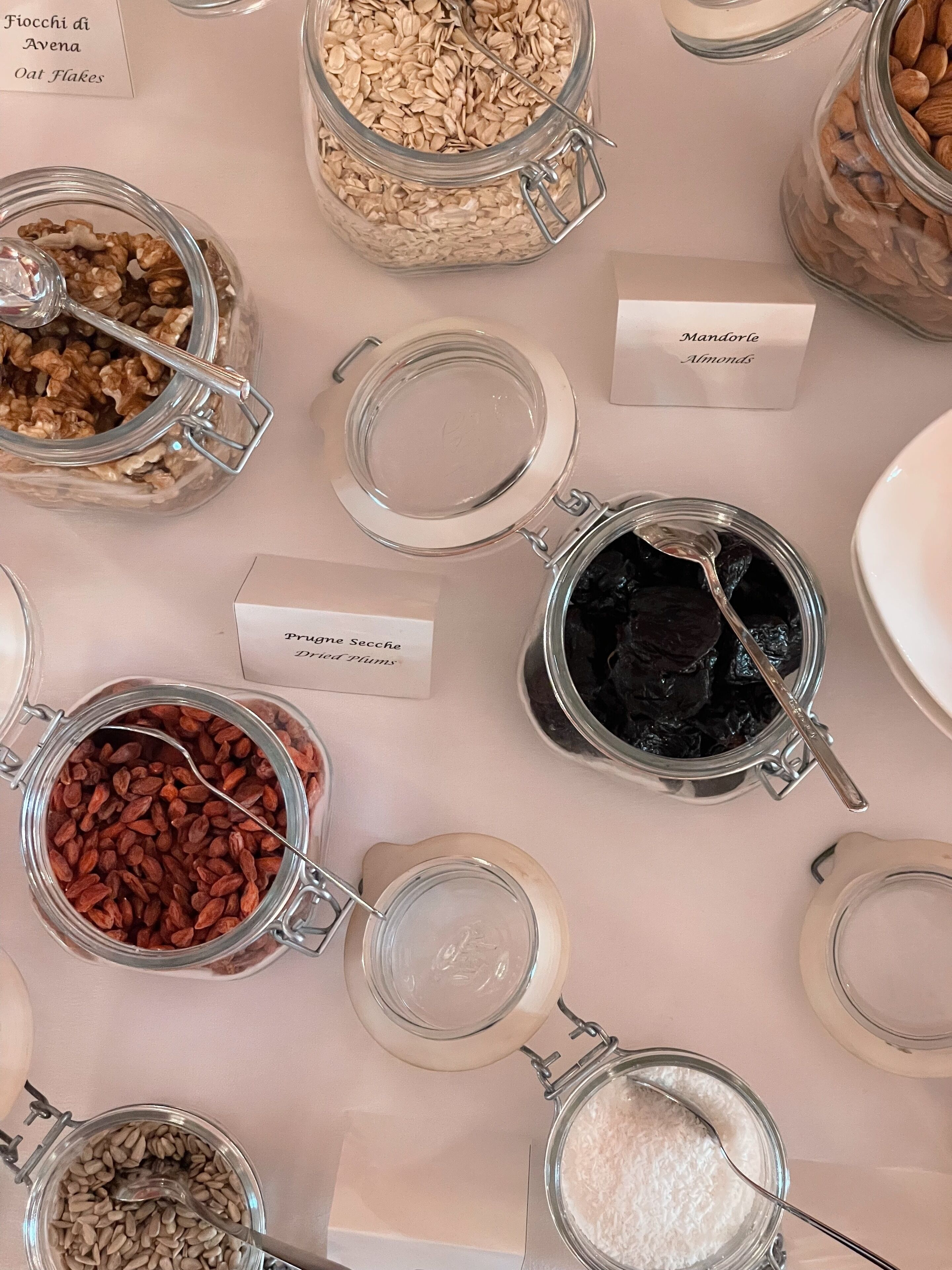 Verschiedene Lebensmittel in Gläsern mit Etiketten, darunter Haferflocken, Mandeln, Dörrpflaumen und andere Nüsse.