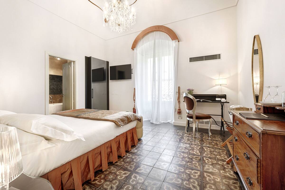 A hotel room with a bed, a desk, a chair, a dresser, a TV, and a chandelier.