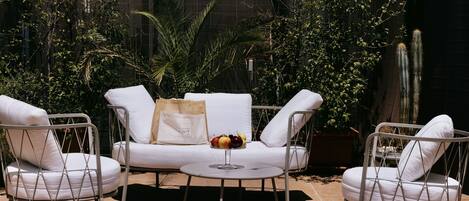 Un patio con un sofá blanco, dos sillas blancas y una mesita redonda con fruteros.