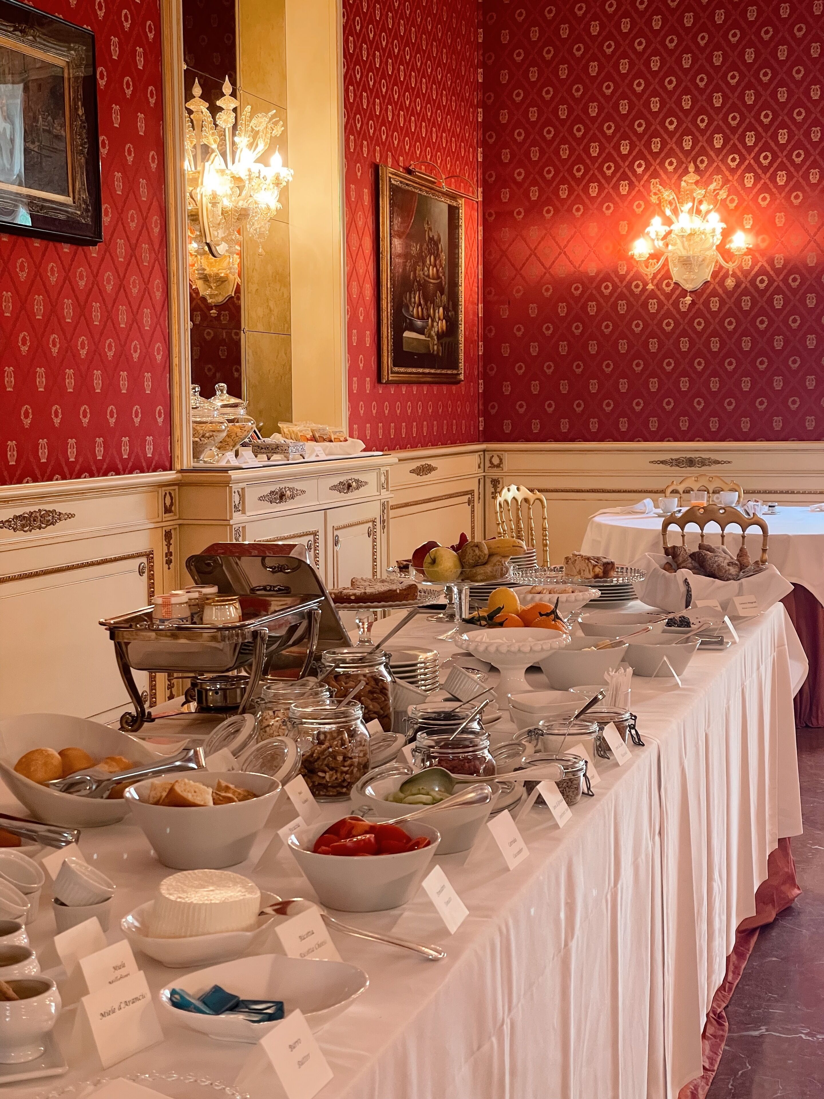 A buffet setup with various food items and utensils on a table, set against a red patterned wall with a chandelier.