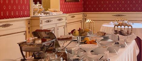 A buffet setup with various food items and utensils on a table, set against a red patterned wall with a chandelier.