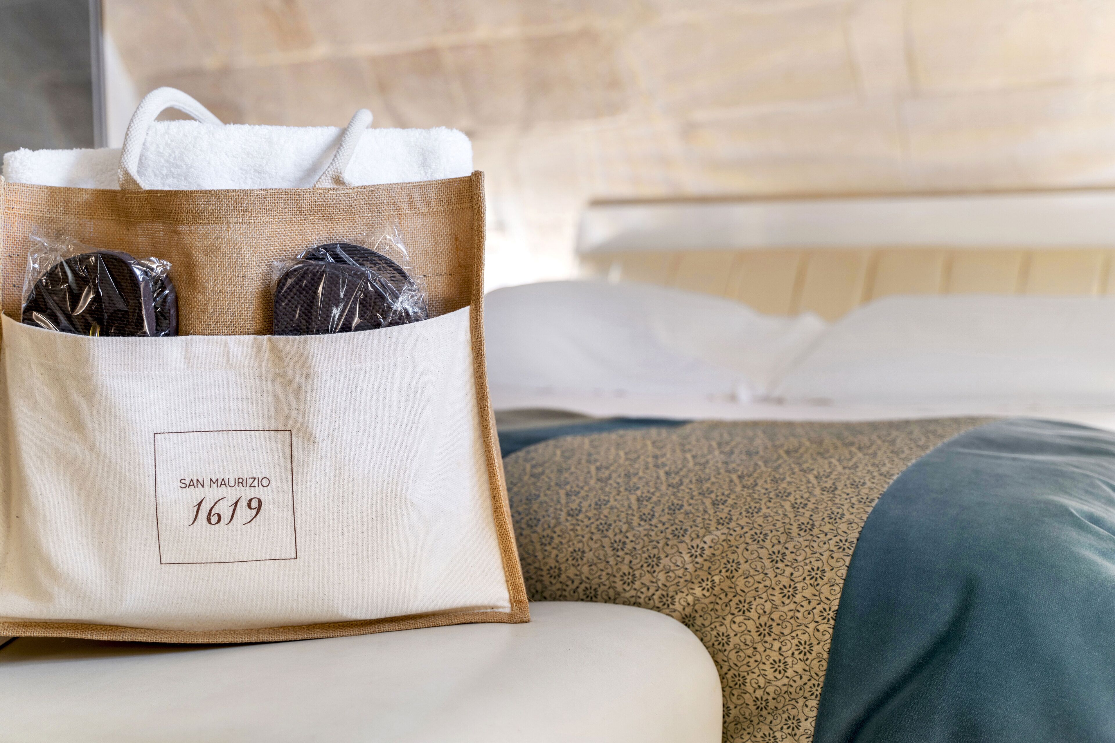 A hotel room with a bed, a pillow, and a bag with the text "SAN MAURIZIO 1619".