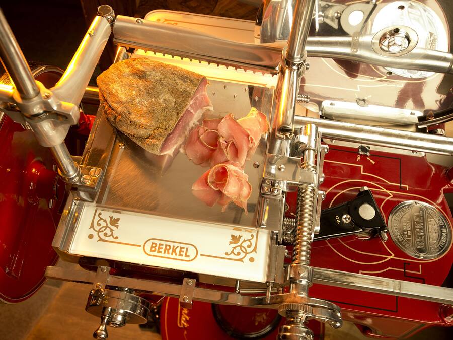 Un cortador de fiambres Berkel rojo con un trozo grande de carne y jamón en lonchas finas.