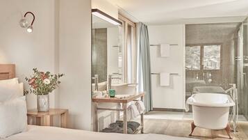 Ein modernes Hotelzimmer mit einem großen Bett, einem Nachttisch mit Blumenvase, einem Badezimmer mit Badewanne und einem großen Spiegel.