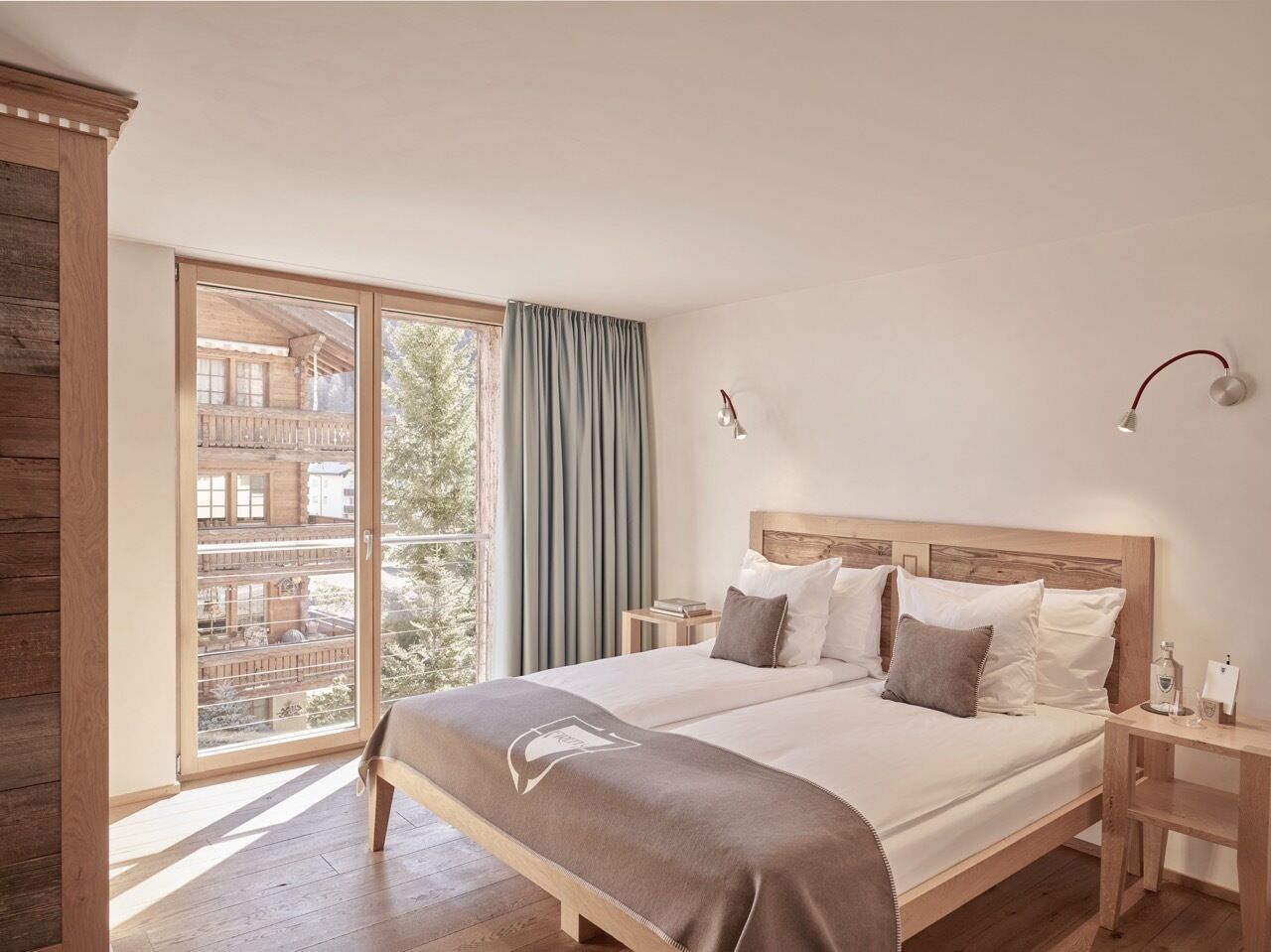 Un dormitorio con una cama grande, cabecera de madera, dos mesitas de noche y vistas a un edificio a través de un ventanal corredera.