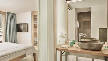 A hotel room with a bed, a wooden nightstand, a large mirror, a stone sink, and a window with curtains.