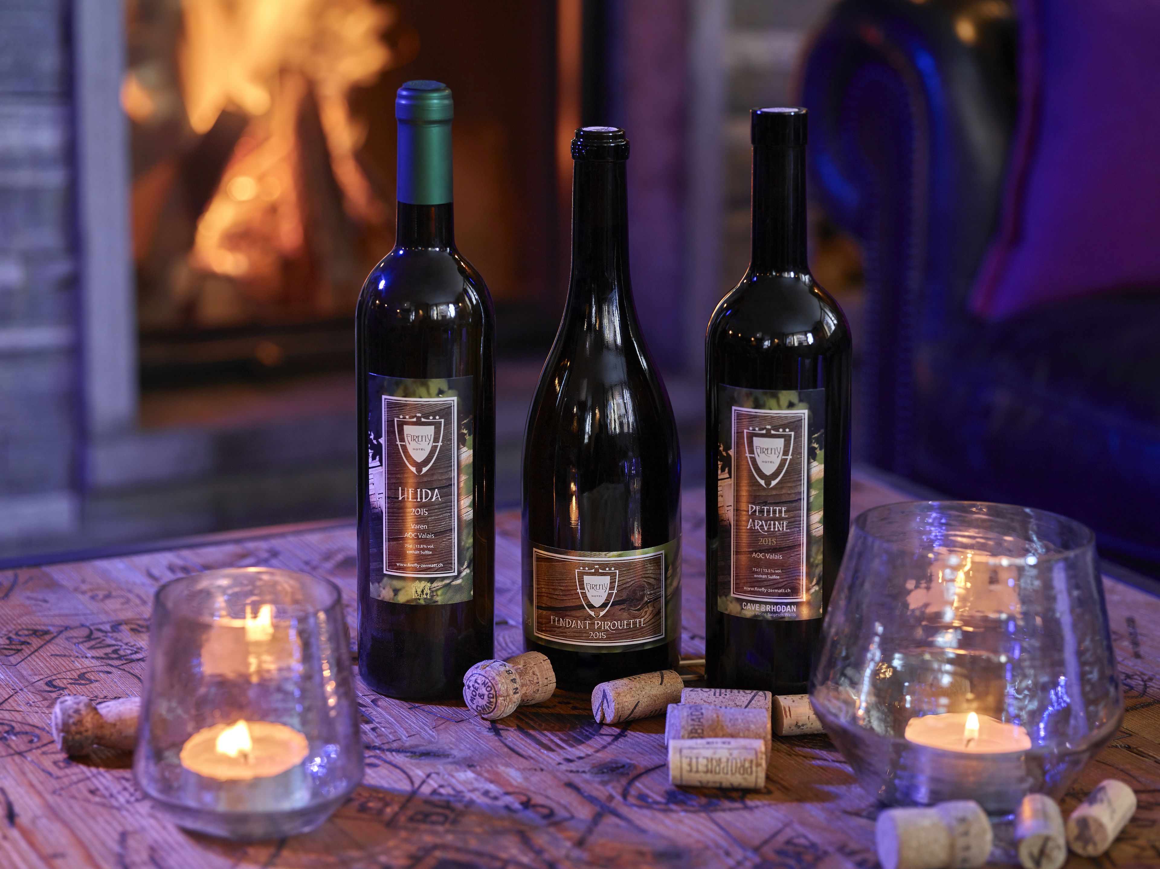 Três garrafas de vinho e duas velas acesas sobre uma mesa de madeira.