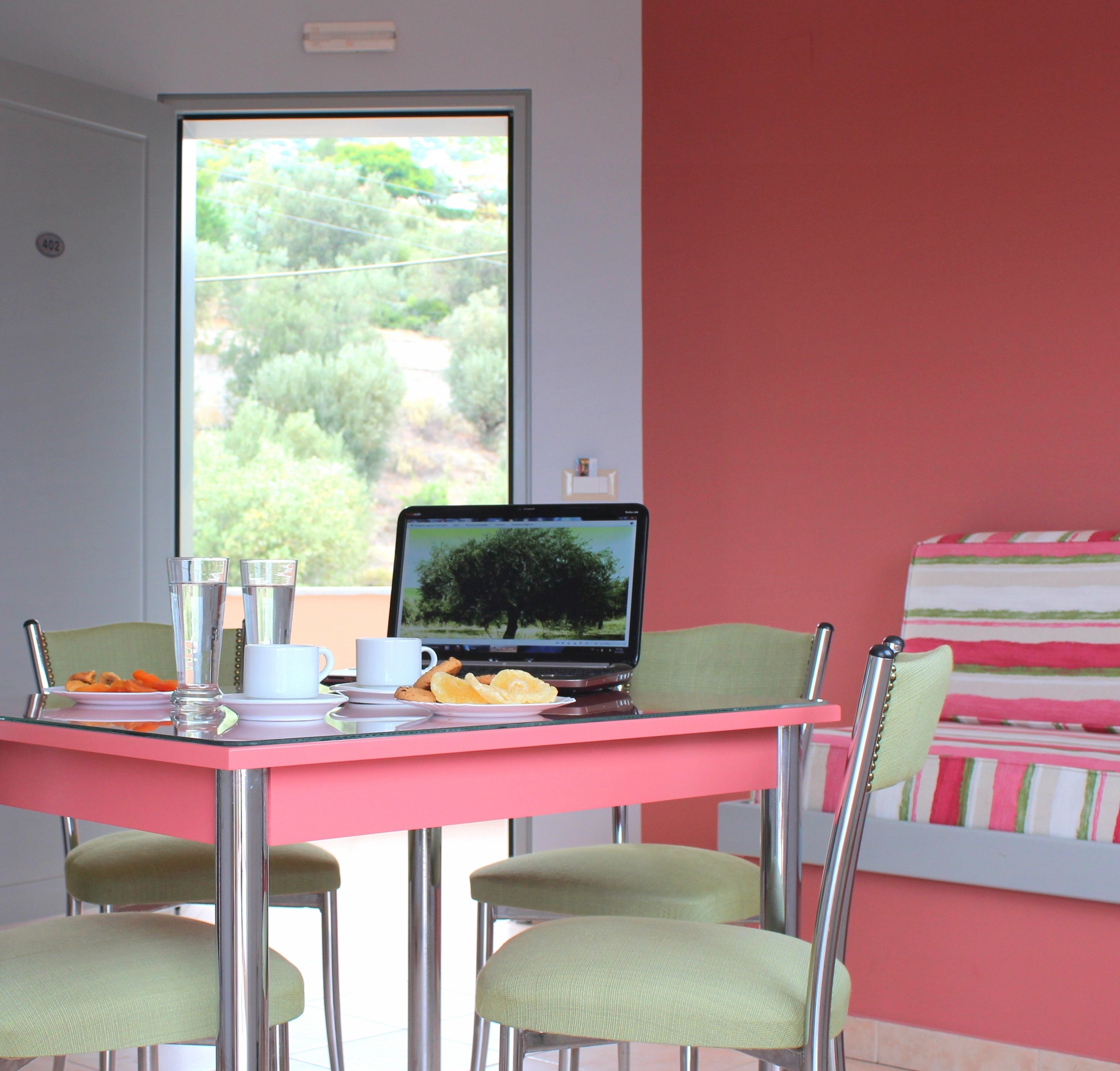 Uma área de refeições com um laptop sobre uma mesa rosa, rodeada por cadeiras e pratos de comida.