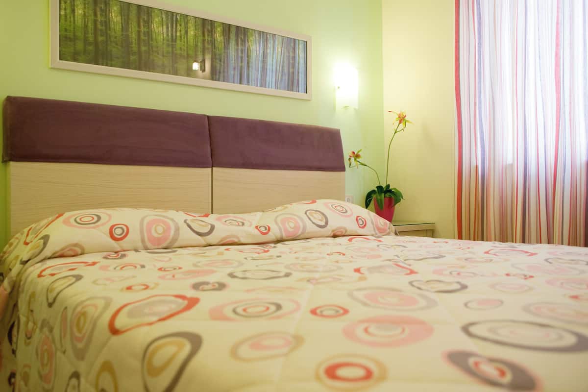 Ein Schlafzimmer mit einem Bett, einem Kopfteil, einem an der Wand hängenden Bild, einem Nachttisch, einer Zimmerpflanze und einem Fenster mit Vorhängen.