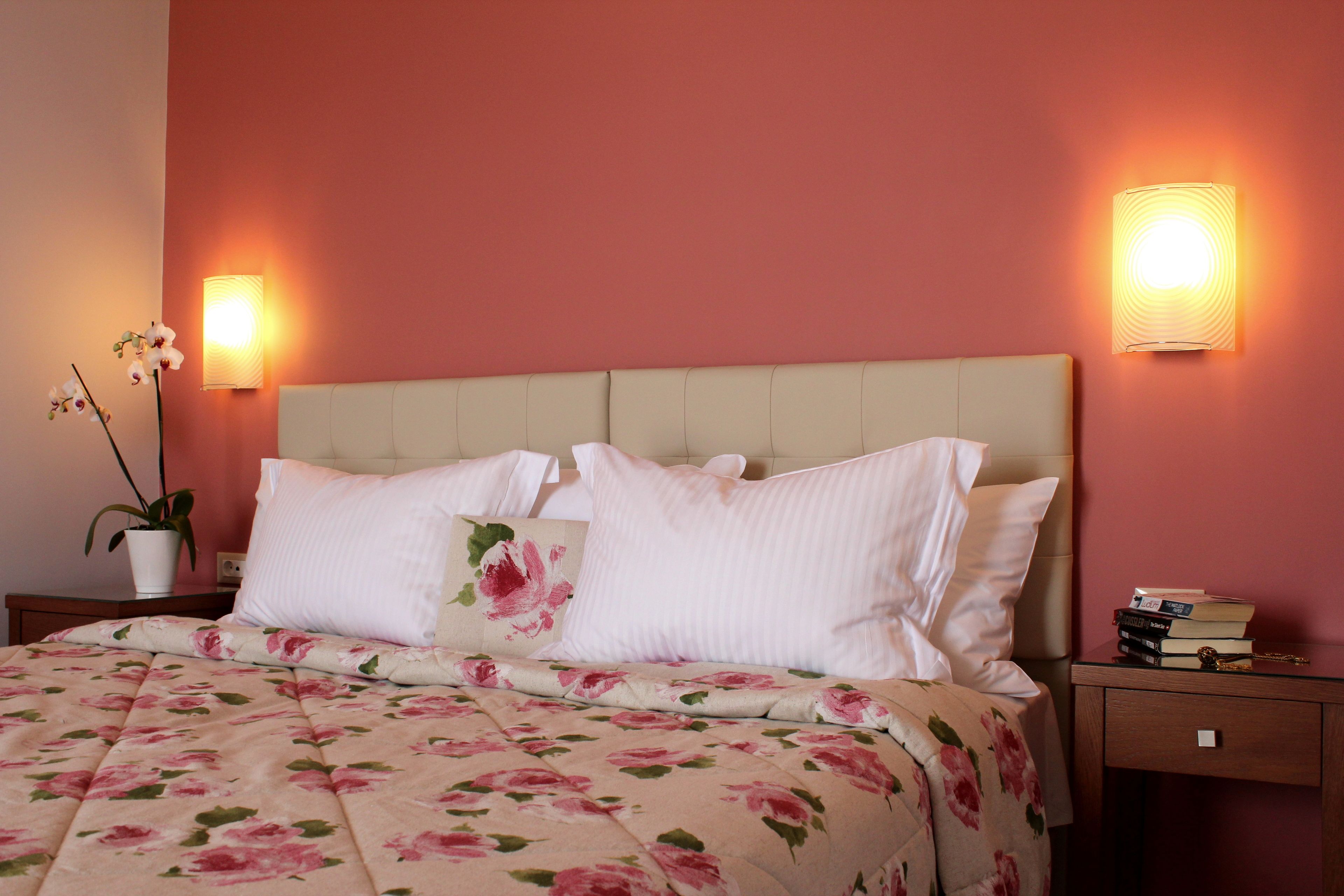Een opgemaakt bed met een bloemenprint, twee bedlampen, een nachtkastje met boeken en een potplant.